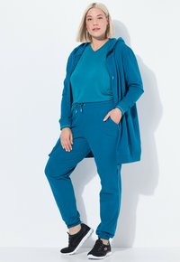 Teal athleisure-set bestående av en zip-up hoodie och joggers med snörning, tillverkad av mjukt tyg. Kombinerat med svarta sportskor.