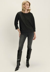 Mujer con cabello rubio que lleva una blusa negra de manga larga texturizada, jeans ajustados grises y botas negras de tacón alto hasta la rodilla, de pie de lado.