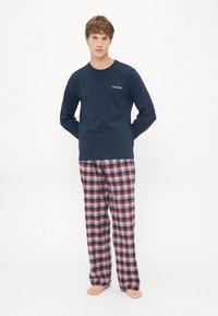 Hombre de pie descalzo, vistiendo una camisa de manga larga azul marino con el logo "GUESS" y pantalones de cuadros rojos, blancos y azules, sobre un fondo blanco liso.