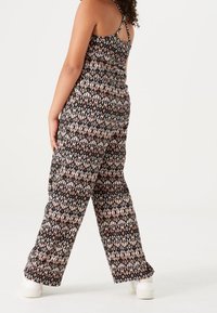 Kobieta w bezrękawowym kombinezonie z czarno-biało-różowym wzorem w romby, stojąca tyłem, widoczne białe buty.