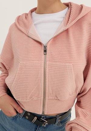 Hoodie zippé côtelé rose clair avec poches avant, porté sur une chemise blanche, associé à un jean bleu et une ceinture noire.