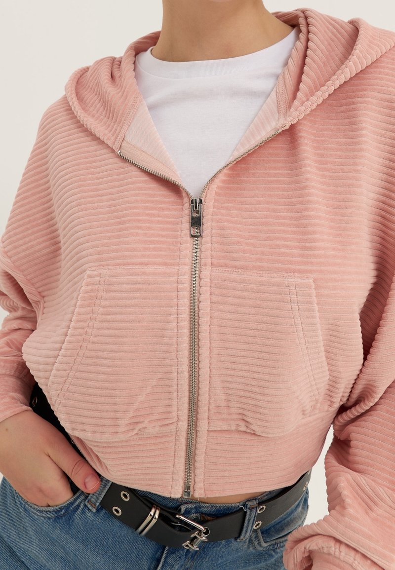Hoodie zippé côtelé rose clair avec poches avant, porté sur une chemise blanche, associé à un jean bleu et une ceinture noire.