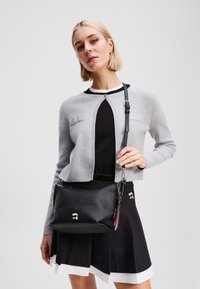 Geantă de umăr din piele neagră cu suprafață texturată, având un accent cu logo și accesorii din argint. Purtați-o cu un cardigan gri și o fustă neagră.