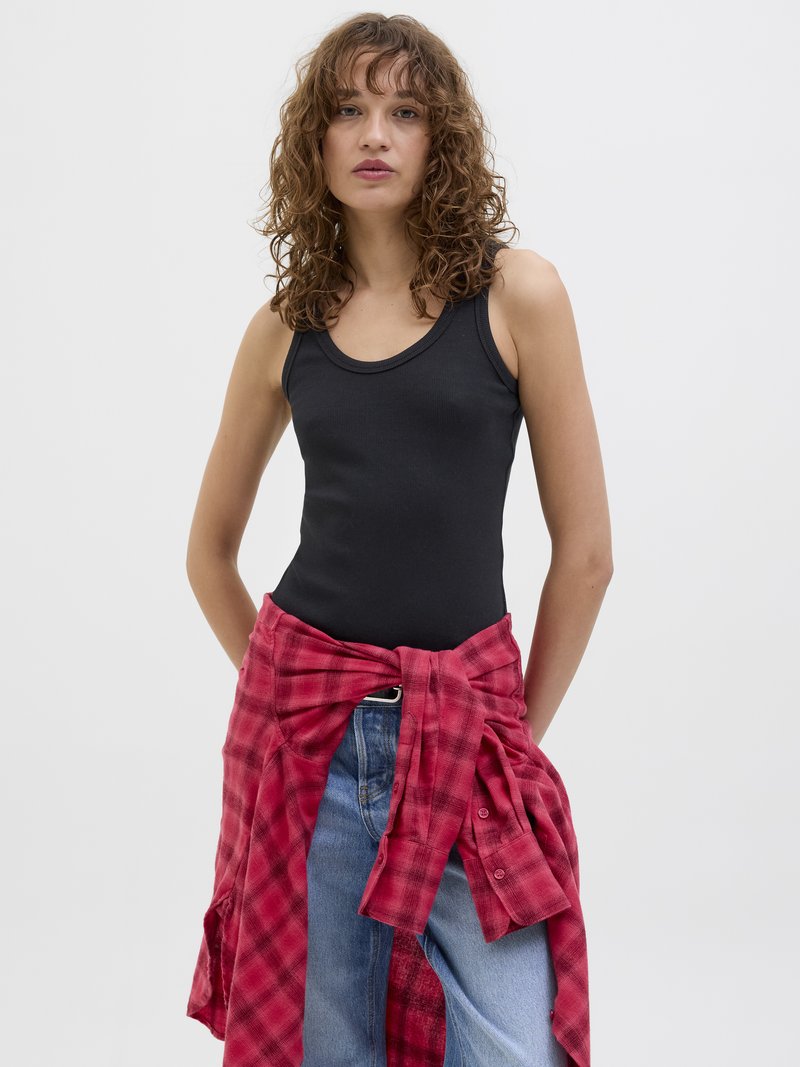Top preto sem mangas com uma textura canelada, combinado com jeans azul claro e uma camisa xadrez vermelha atada à cintura.
