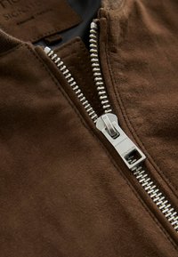 Chaqueta de ante marrón con un zipper metálico plateado. Primer plano del zipper, mostrando los dientes y la lengüeta, con una textura suave alrededor de la abertura.