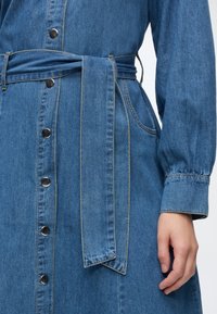 Μπλε φόρεμα από denim με κουμπιά μπροστά, ταιριαστή υφασμάτινη ζώνη δεμένη στη μέση, πλαϊνή τσέπη και μακρύ μανίκι με μανσέτα στο δεξί χέρι.