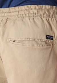 Beżowe spodnie z elastycznym paskiem, z małą kieszonką z tyłu i czarną metką marki "JACK & JONES". Gładka tekstura.