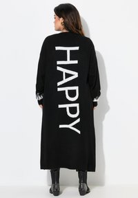 Cardigan en maille noire avec un grand texte blanc "HAPPY" au dos, des poignets côtelés et des détails complexes aux manches.