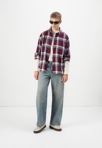 Ung person iført sorte solbriller, en bordeaux skotsk ternet skjorte over en hvid langærmet top, løse blå jeans og beige sneakers, stående mod en hvid baggrund.