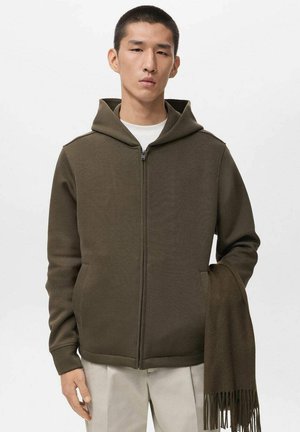 Sudadera con cremallera de color verde oliva, con capucha forrada, bolsillos laterales y puños de canalé, acompañada de una bufanda a juego de color oliva colgada de un brazo.