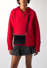 Suéter de punto rojo con cuello tipo polo y puños acanalados, combinado con una falda plisada a cuadros grises. Un pequeño bolso cruzado negro presenta una parte delantera texturizada.