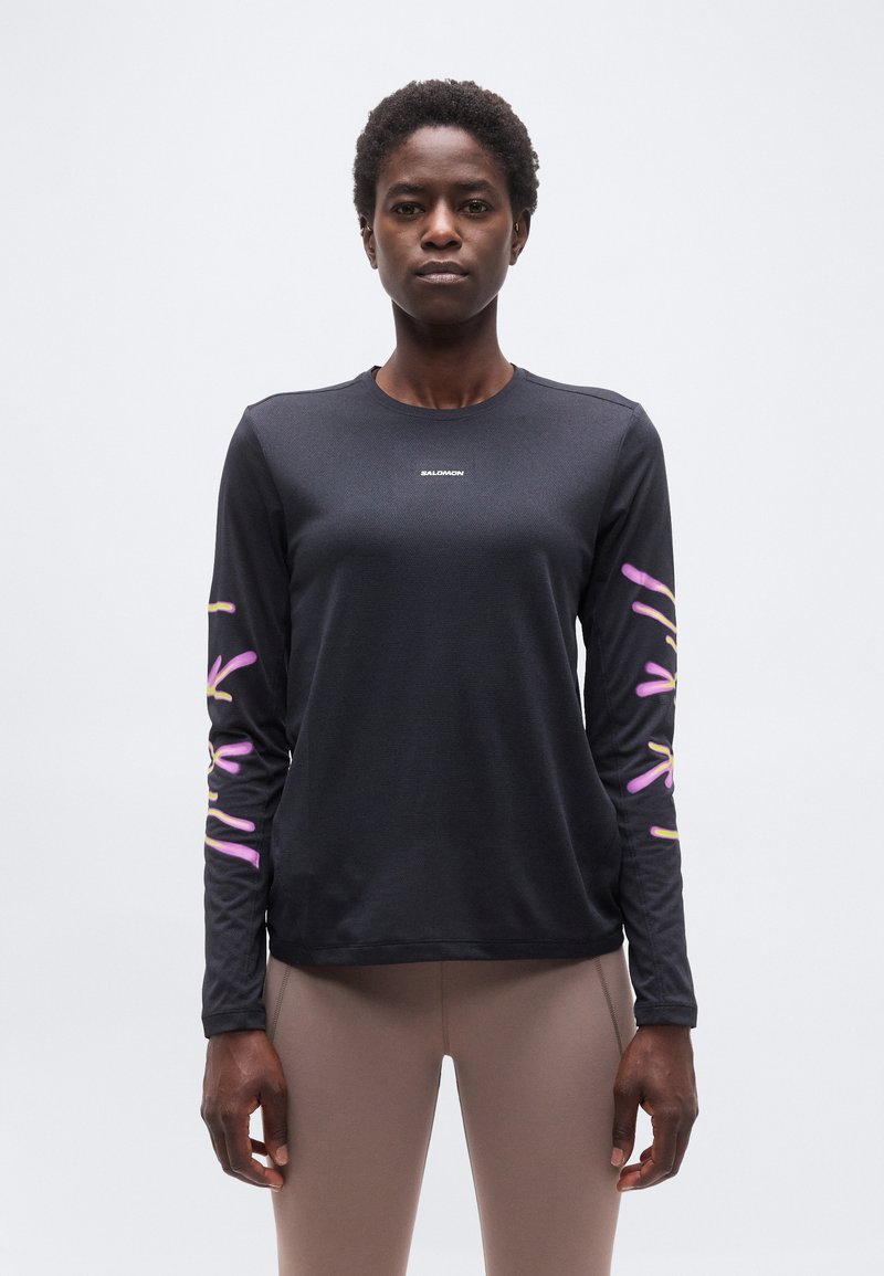 Γυναίκα με μαύρο μπλουζάκι μακρυμάνικο με ροζ και κίτρινα λουλουδένια σχέδια στα μανίκια και μπεζ κολάν, στέκεται μπροστά σε απλό φόντο.