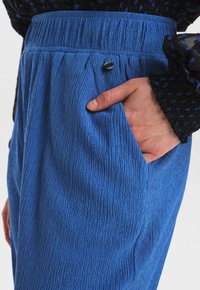 Zbliżenie ręki w kieszeni teksturowanych, niebieskich spodni z wysokim stanem z czarnym guzikiem, w połączeniu z rękawem przezroczystej bluzki w czarne i niebieskie kropki.