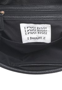 Interno di una borsa nera con un'etichetta di tessuto che mostra il testo ripetuto: "DEVO RESISTERE" seguito da "L'HO COMPRATA." La fodera è nera tinta unita.