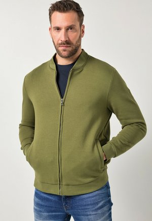 Uomo con barba che indossa una giacca verde oliva con cerniera e jeans blu, in piedi con le mani nelle tasche della giacca contro uno sfondo semplice.