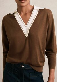 Maglione marrone a collo a V con colletto bianco e bordi a contrasto. Tessuto morbido e maniche lunghe, abbinato a jeans blu scuro.