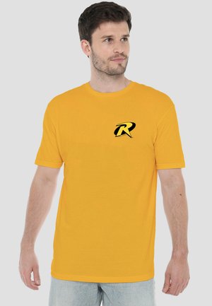 Mann trägt ein gelbes T-Shirt mit kurzen Ärmeln und einem schwarzen und gelben "R"-Logo auf der linken Brust, steht vor einfachem grauem Hintergrund.