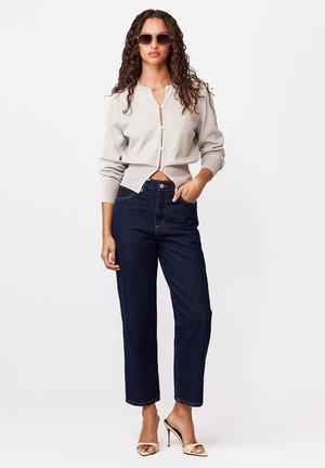 Kobieta o kręconych włosach, w okularach przeciwsłonecznych, lekkim rozpinanym swetrze, ciemnych jeansach o prostych nogawkach i beżowych sandałach na wysokim obcasie, stoi z jedną ręką w kieszeni.