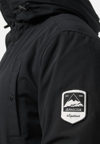 Black hooded jacket featuring a water-resistant material, zippered front, chest pocket, and a patch with mountain logo and text on the sleeve.