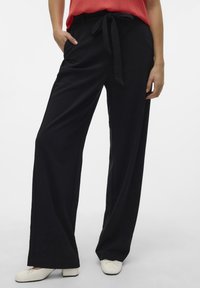 Pantaloni neri a gamba larga con una texture liscia, dotati di vita legata e tasche laterali. Indossati con scarpe basse bianche.