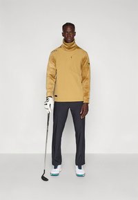 Haut de golf beige avec manches texturées, poche zippée, associé à un pantalon foncé et des chaussures de golf. Le modèle tient un club de golf, debout sur un fond neutre.