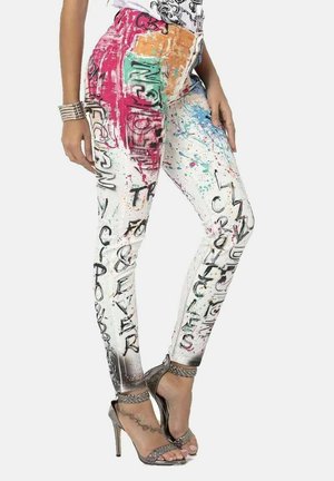 Woman wearing colorful graffiti-style skinny jeans and silver high-heeled sandals with ankle straps, standing with one hand resting on hip.
