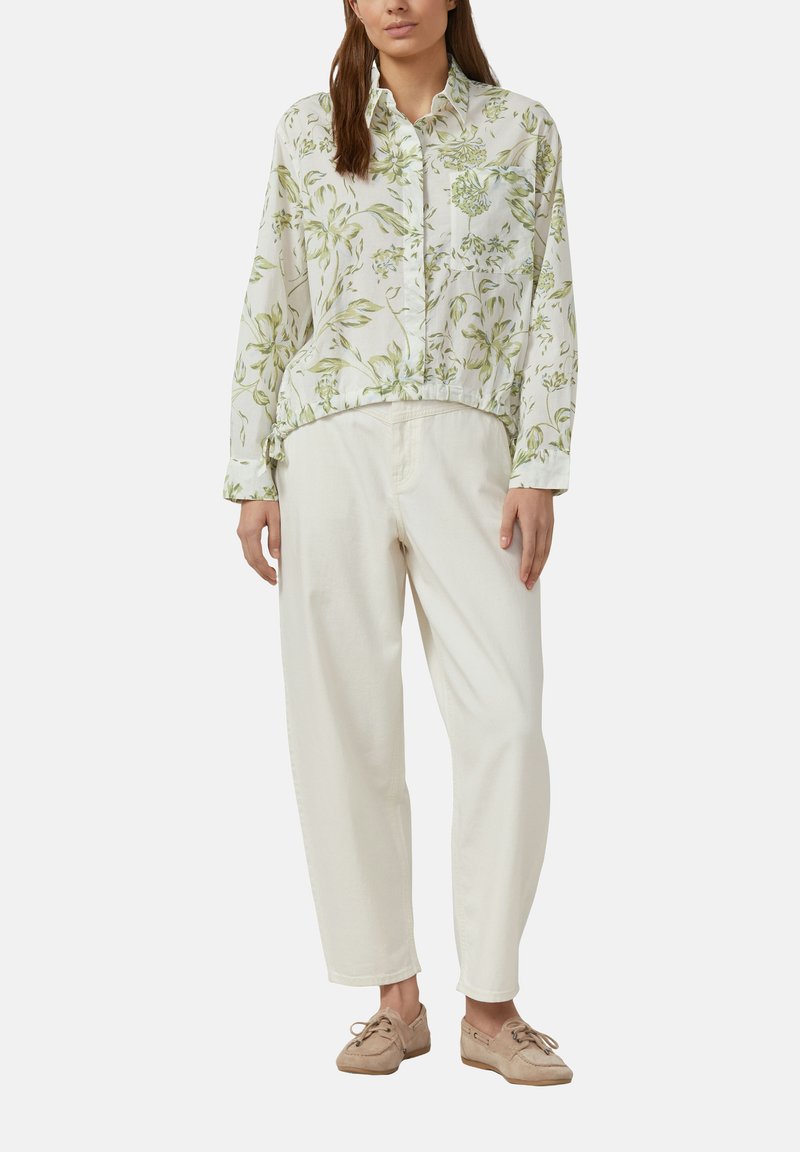 Frau trägt ein weißes, langärmliges Hemd mit Blumenmuster, locker sitzende cremefarbene Hose und beige Schnür-Loafer, steht.