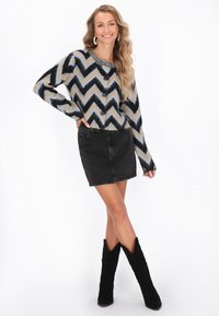 Minirock aus schwarzem Denim, kombiniert mit einer chevron-musterierten Strickjacke in Blau, Schwarz und Beige. Die Strickjacke hat Knöpfe und lange Ärmel. Schwarze kniehohe Stiefel.