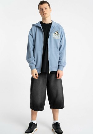 Młody mężczyzna stojący, ubrany w jasnoniebieską bluzę na zamek z grafiką, czarne, oversize spodenki do kolan, czarne trampki i białe skarpetki.