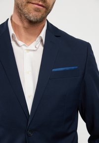 Sötétkék blézer sima textúrával, egy gombbal, és kék zsebkendővel. Fehér ing látható gombokkal alatta.