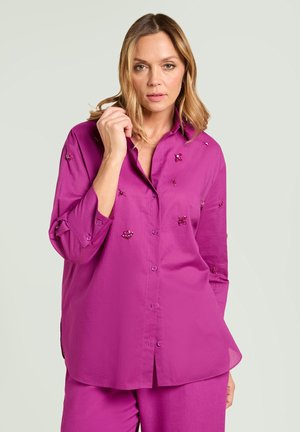 Donna che indossa una camicetta viola brillante con bottoni e decorazioni di perline e pantaloni abbinati, in piedi davanti a uno sfondo chiaro e semplice.
