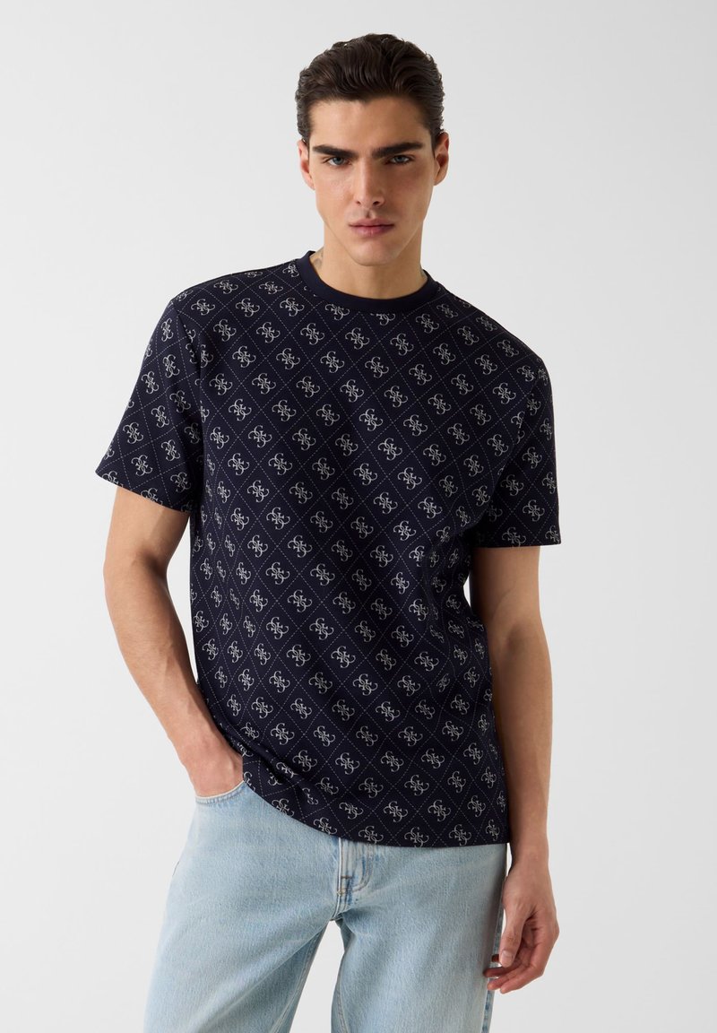 Νέος άνδρας που φοράει μπλε σκούρο μπλουζάκι με σχέδια και ανοιχτόχρωμα μπλε τζιν, ποζάροντας με το ένα χέρι στην τσέπη, μπροστά σε απλό φόντο.