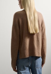 Frau mit langen blonden Haaren, die einen braunen Pullover und bluejeans trägt, mit dem Rücken zu einer schlichten weißen Wand.