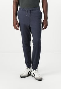 Navy jogger pants with an elastic waistband and cuffs, featuring a smooth texture and drawstring detail, paired with grey and white sneakers.