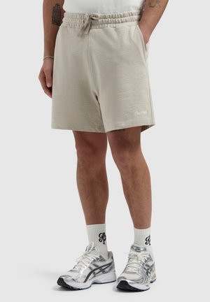 Onderlichaam van een persoon die beige short met trekkoord draagt, witte merksokken en zilver-witte hardloopschoenen op een effen achtergrond.