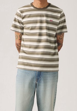 Mand iført olivengrøn og hvid stribet Levi's t-shirt og lyseblå jeans med synlige tatoveringer på begge underarme.