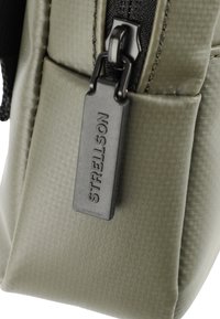 Zelena teksturirana platnena torba z črnim zadrgo. Ima pravokotno obliko in oznako logotipa Strellson, pritrjeno na pull zadrgo.