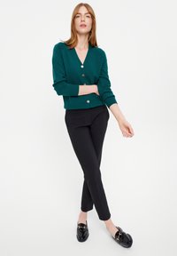Cardigan verde con bottoni, maniche lunghe, scollatura a V e bottoni dorati, abbinato a pantaloni neri aderenti e mocassini neri con dettagli dorati.