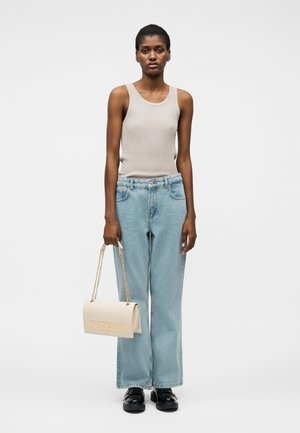 Kvinde iført en beige tank top, lyseblå jeans med vide ben, sorte sko, der holder en beige Love Moschino håndtaske med guldkæde.