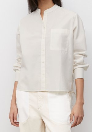 Ljusbeige skjorta med mandarin-krage, knappar fram och en platt ficka på vänster sida. Tillverkad av slät tyg med avslappnad passform.