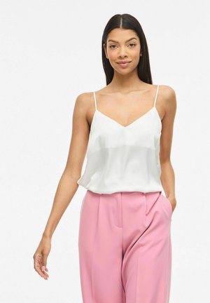 Kvinde med langt mørkt hår iført en hvid ærmeløs top og lyserøde bukser med høj talje, stående med den ene hånd i lommen.