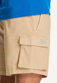 Shorts cargo bej dintr-un material ușor, având un buzunar lateral, textură netedă și un design minimal. Persoana care le poartă are o cămașă albastră.