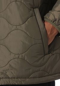 Handen i fickan på en olivgrön vadderad jacka med vågigt sömnad och elastisk mudd på ärmen.