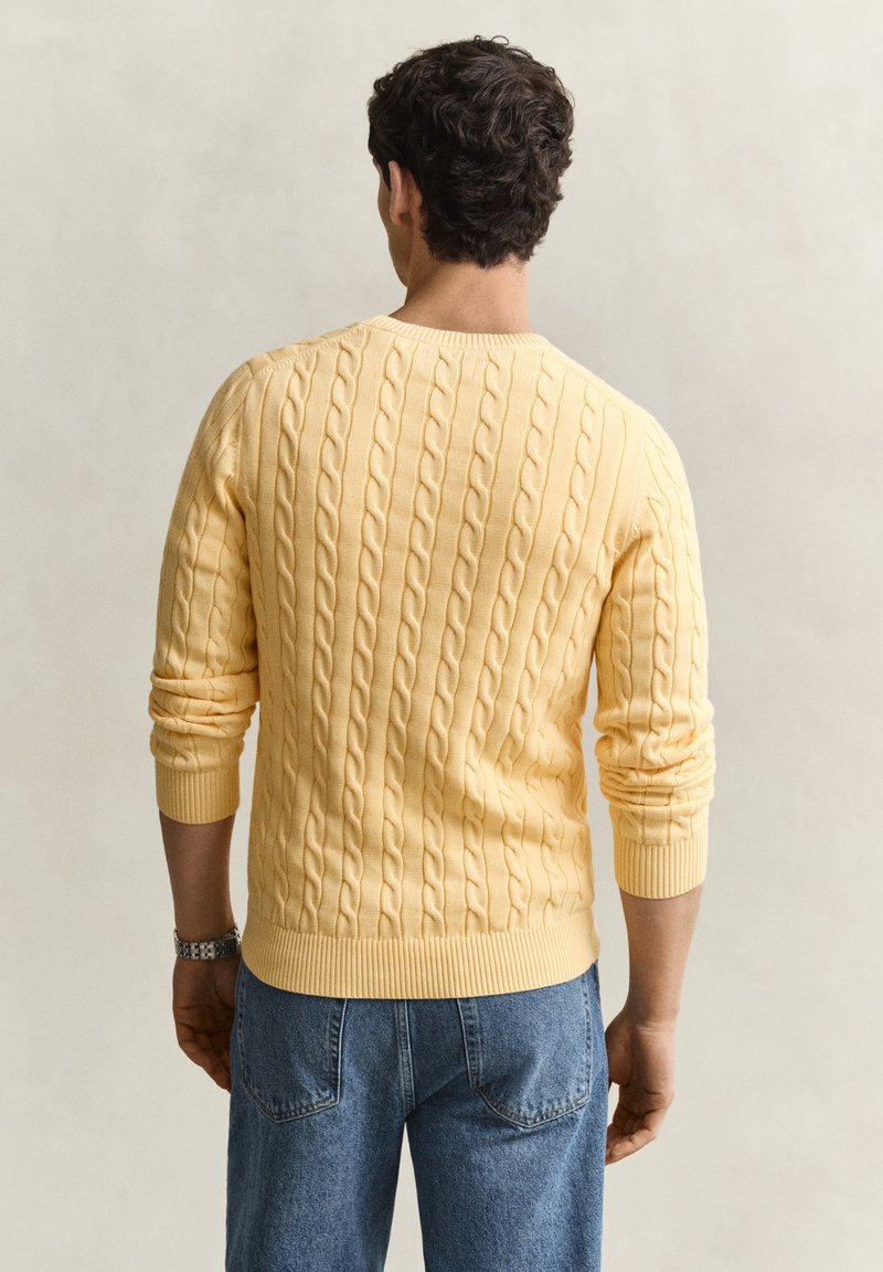 Homme aux cheveux bouclés foncés portant un pull jaune torsadé et un jean bleu, debout, dos à un fond clair uni.