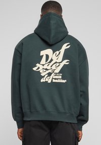 Mørkegrønn hettegenser med en stor kremfarget grafikk som viser ordet "Def" i stilisert tekst på baksiden. Har en foret hette og oppbrettede ermer.