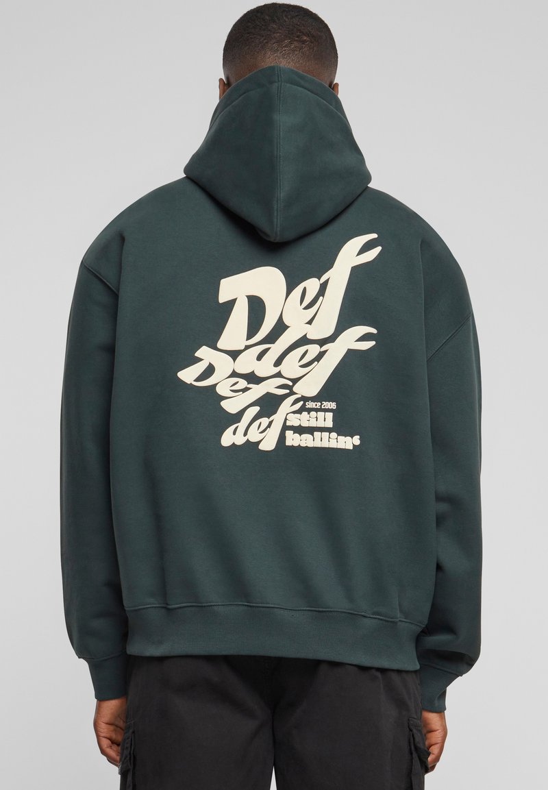 Mørkegrønn hettegenser med en stor kremfarget grafikk som viser ordet "Def" i stilisert tekst på baksiden. Har en foret hette og oppbrettede ermer.