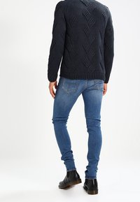 Dunkel gestrickter Pullover mit strukturiertem Rautenmuster, kombiniert mit blauen Slim-Fit-Jeans und schwarzen Leder-Ankle-Boots.