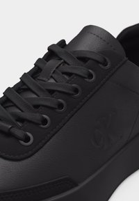 Κοντινό πλάνο μαύρου δερμάτινου αθλητικού παπουτσιού με κορδόνια στον ίδιο τόνο, μεταλλικά χαντζάκια και διακριτικό ανάγλυφο λογότυπο "CK" στην πλευρική επιφάνεια.