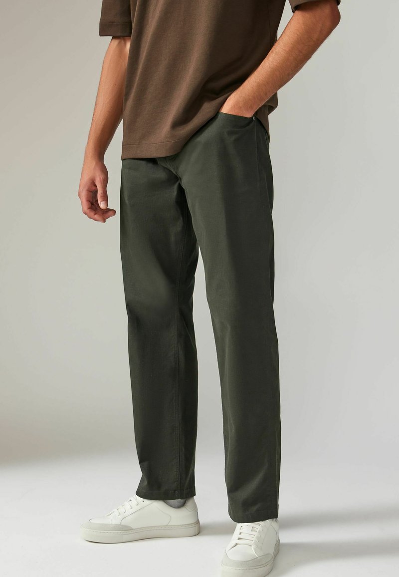 Chino droits vert olive en coton, avec poches latérales, portés avec des baskets blanches. L'arrière-plan est gris neutre.