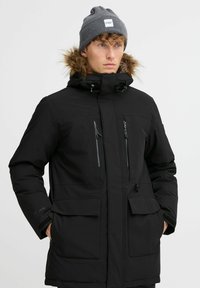Crna izolirana parka jakna s kapuljačom obloženom krznom, zatvorenim džepovima na prsima i prednjim zatvaranjem na dva dugmeta. Model nosi sivi bejzbolac.